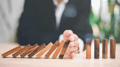 Das Bild zeigt Dominosteine, die in einer Reihe stehen. Von links kippen die Dominosteine nacheinander um; eine Hand in der Mitte der Reihe stoppt die Steine und verhindert, dass die Steine auf der rechten Seite ebenfalls umkippen. Das Bild soll zeigen, dass durch rechtzeitige Intervention und Präventionsmaßnahmen, die weitere Eskalation eines Konflikts gestoppt werden kann.