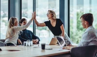 Das Bild ein Team von vier Mitarbeitenden (zwei Frauen und zwei Männer) an einem Besprechungstisch in einem modernen Büro aus Glas. Drei Mitglieder sitzen am Tisch, eine Frau steht und klatscht der anderen, sitzenden Frau euphorisch in die Hand. Das Bild soll zeigen, dass das Team durch gute Zusammenarbeit und gute Stimmung einen Erfolg erzielt hat.