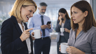 Mobbing im Büroalltag: Eine Kollegin spricht eine jüngere Mitarbeiterin mit spöttischem, herabwürdigendem Ton an, während diese verunsichert reagiert. Zwei weitere Personen im Hintergrund schauen weg. Szene verdeutlicht: Mobbing und Führung -Warum Wegschauen Mobbing begünstigt.