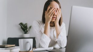 Eine erschöpfte Frau sitzt an ihrem Schreibtisch, stützt ihr Gesicht in die Hände und wirkt gestresst. Das Bild symbolisiert den Arbeitsdruck und die Herausforderungen für das Wohlbefinden und die psychische Gesundheit von Frauen am Arbeitsplatz