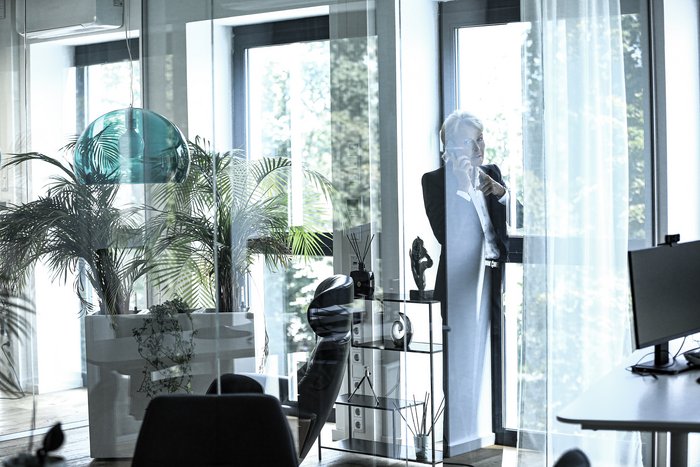 Das Bild zeigt Beate Reinhart in einem modernen Büro mit viel Glas, moderner Einrichtung und Palmen als Raumbegrünung. Beate Reinhart im dunklen Businessanzug steht am Fenster, sie telefoniert mit dem Mobiltelefon und dreht sich dabei freundlich in den Raum blickend um.   