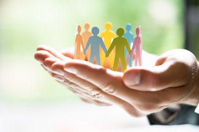 Das Bild zeigt zwei aneinander liegende Hände, deren Innenflächen nach oben zeigen. Die Hände halten einen Ring von verschieden farbigen Männchen. Diese sollen ein diverses Team darstellen, die von den Händen als Symbol für die Unternehmenskultur, schützend umfasst werden.